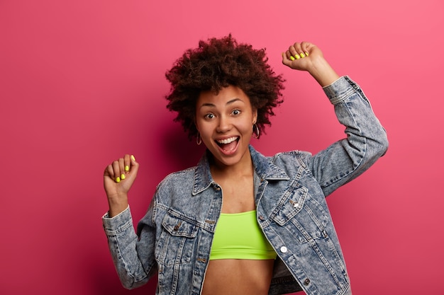 Retrato de uma linda adolescente dança feliz, mantém os braços levantados e fecha os punhos, gosta de boa música na festa, usa blusa e jaqueta jeans, isolada na parede rosa, sente-se otimista e despreocupada