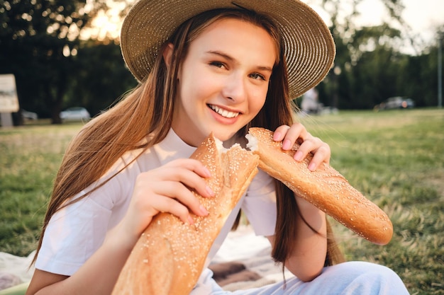 Retrato de uma linda adolescente sorridente com chapéu de palha segurando pão baguete alegremente olhando para a câmera no piquenique no parque da cidade