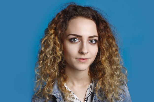 Retrato de uma linda aluna com a pele limpa e perfeita e cabelos claros ondulados posando, vestida com uma jaqueta jeans contra uma parede azul em branco com espaço de cópia para seu conteúdo de publicidade