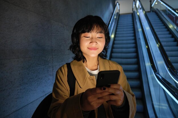 Retrato de uma linda e feliz garota asiática parada com smartphone na escada rolante descendo para a estação de metrô