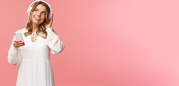 Foto grátis retrato de uma linda e linda garota caucasiana loira em vestido branco sonhando acordado enquanto escuta favorit