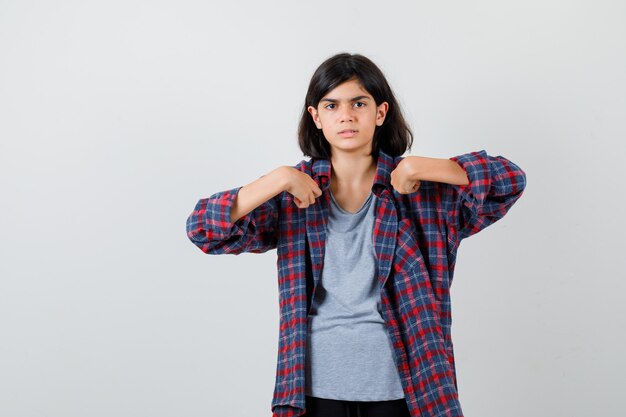 Retrato de uma linda garota adolescente apontando para si mesma em uma camisa quadriculada e parecendo confusa com a vista frontal