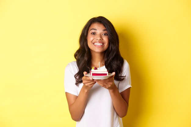 Retrato de uma linda garota afro-americana comemorando aniversário, sorrindo e parecendo feliz, segurando um bolo de aniversário com uma vela, em pé sobre um fundo amarelo