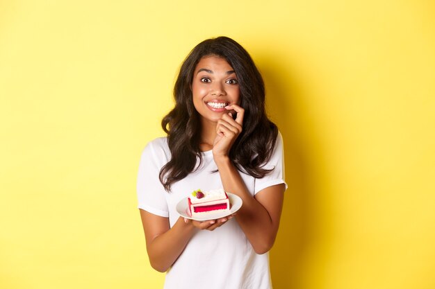 Retrato de uma linda garota afro-americana sorrindo, segurando um delicioso pedaço de bolo, tentada a comer a sobremesa, em pé sobre um fundo amarelo