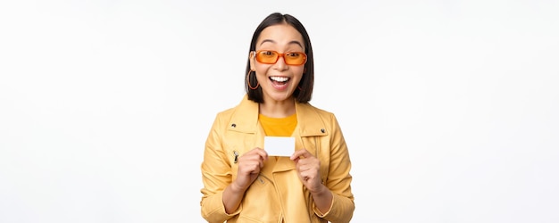 Retrato de uma linda garota asiática moderna em óculos de sol sorrindo feliz mostrando cartão de crédito sobre fundo branco