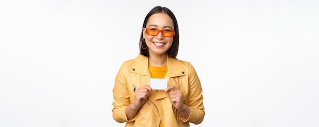 Retrato de uma linda garota asiática moderna em óculos de sol sorrindo feliz mostrando cartão de crédito sobre o espaço de cópia de fundo branco