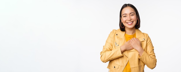 Retrato de uma linda garota asiática sorridente, olhando com apreço, obrigado gesto de mãos dadas no coração lisonjeado em pé sobre fundo branco