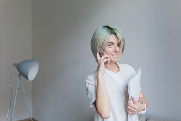 Retrato de uma linda garota com penteado curto cinza andando com papéis em estúdio cinza ... ela usa vestido branco e maquiagem leve. ela está falando ao telefone e sorrindo para a câmera.