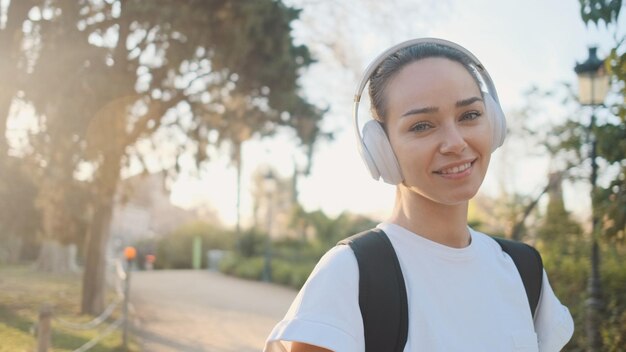 Retrato de uma linda garota em fones de ouvido com mochila sorrindo para a câmera a caminho do treinamento matinal no parque da cidade