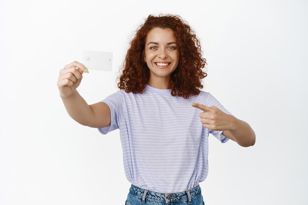 Retrato de uma linda garota encaracolada apontando e segurando o cartão de crédito, torna-se cliente do banco, anunciando descontos, em branco.