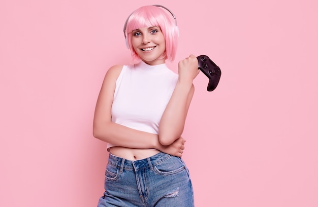 Retrato de uma linda garota gamer feliz com cabelo rosa jogando videogame usando joystick em colorido no estúdio