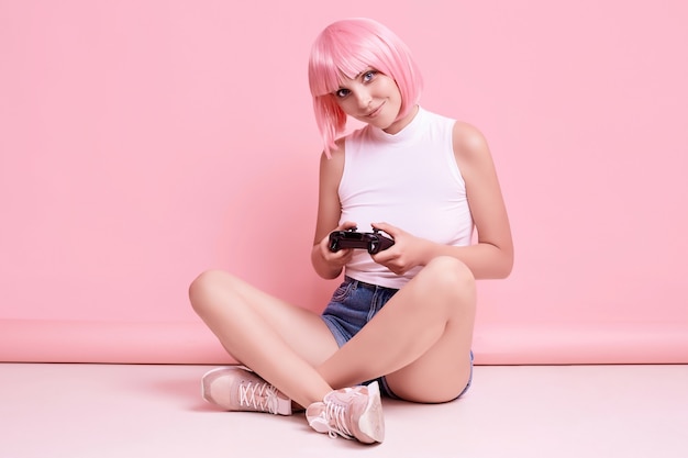 Retrato de uma linda garota gamer feliz com cabelo rosa jogando videogame usando joystick em colorido no estúdio