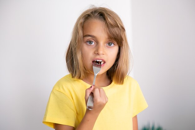 Retrato de uma linda garota loira comendo bolo com um garfo. Criança muito branca em pé, comendo. Conceito de infância, celebração e férias