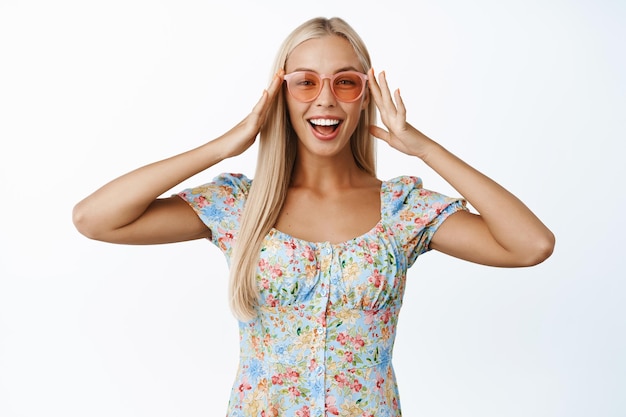 Retrato de uma linda garota loira sorridente em óculos de sol olhando espantado e feliz com a câmera posando com roupa de verão contra fundo branco