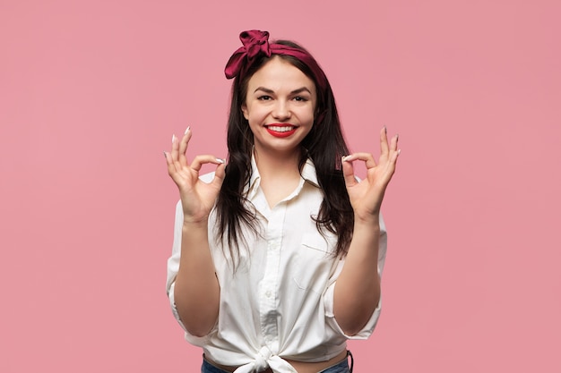 Retrato de uma linda garota pin up vestindo camisa branca e lenço vermelho na cabeça, fazendo gesto de aprovação com as duas mãos