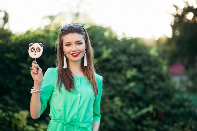 Retrato de uma linda garota sorridente de vestido verde, mostrando na câmera doces como panda na vara na mão. Mulher elegante com lábios vermelhos e brincos longos, segurando a mão no bolso e posando ao ar livre.