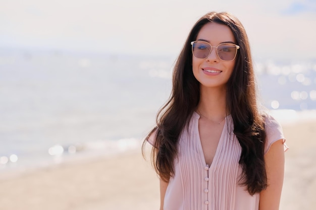 Retrato de uma linda garota sorridente em óculos de sol e vestido rosa na calma à beira-mar.