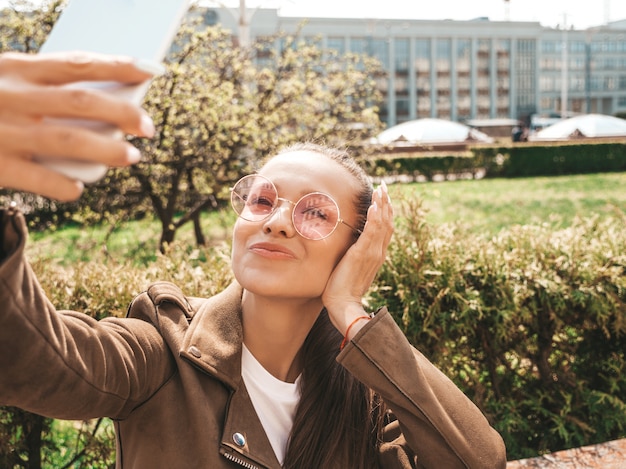 Retrato de uma linda menina morena sorridente no verão hipster jaqueta e calça jeans modelo tomando selfie em smartphone