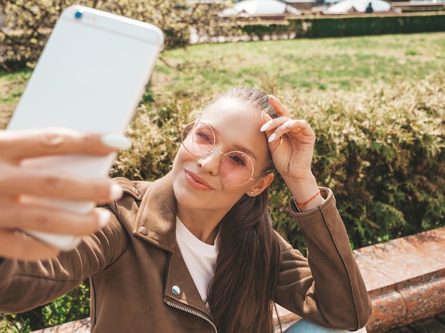 Retrato de uma linda menina morena sorridente no verão hipster jaqueta e calça jeans modelo tomando selfie em smartphone