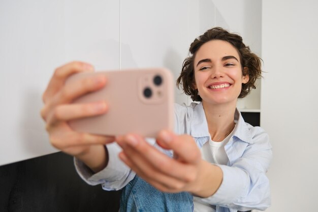 Retrato de uma linda menina morena tira selfie em smartphone em sua cozinha posando para foto em mo