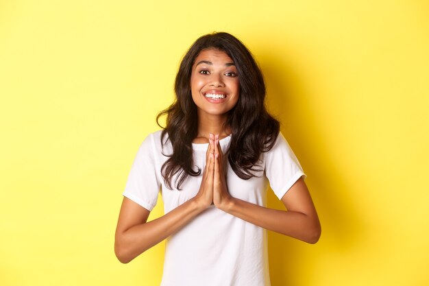 Retrato de uma linda mulher afro-americana, sorrindo e de mãos dadas para rezar, dizendo obrigado, sentindo-se grato, de pé sobre fundo amarelo.