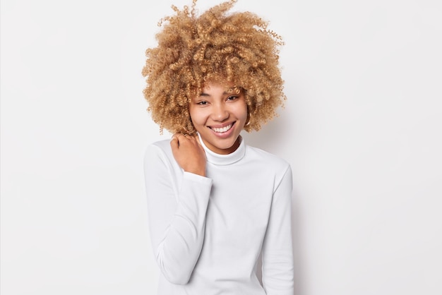 Foto grátis retrato de uma linda mulher alegre com cabelo espesso encaracolado mantém a mão no ombro sorri agradavelmente usa gola rulê casual gosta de bom dia tem uma conversa agradável com poses de interlocutor no interior