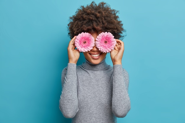 Retrato de uma linda mulher alegre com uma expressão feliz, tem beleza natural, cobre os olhos com margarida gerbera rosa, vestida em poses casuais de gola alta cinza