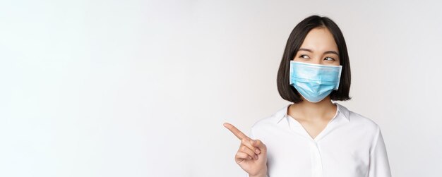 Retrato de uma linda mulher asiática com máscara facial médica proteção contra coronavírus apontando o dedo para a esquerda e lo
