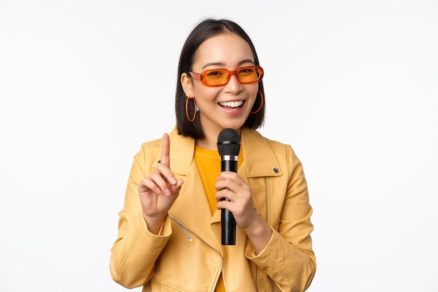 Retrato de uma linda mulher asiática em óculos de sol menina elegante cantando dando discurso com microfone segurando o microfone e sorrindo em pé sobre fundo branco Copie o espaço