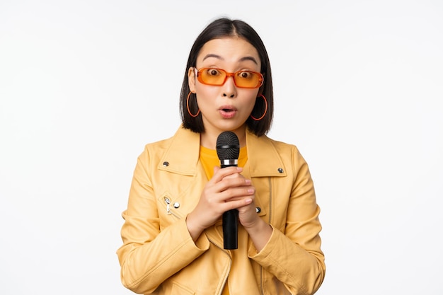 Retrato de uma linda mulher asiática em óculos de sol menina estilosa cantando dando discurso com microfone segurando microfone e sorrindo em pé sobre fundo branco