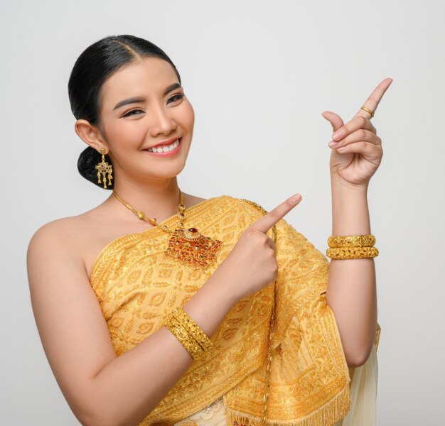 Retrato de uma linda mulher asiática em traje de vestido tradicional tailandês sorri e aponta o dedo com uma pose graciosa na parede branca