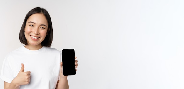 Retrato de uma linda mulher asiática mostrando o polegar para cima e a tela do smartphone sorrindo enquanto anunciava m