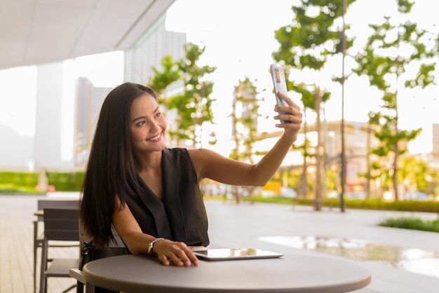 Retrato de uma linda mulher asiática sentada ao ar livre em um restaurante cafeteria, tirando uma selfie com o celular