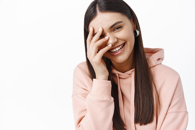 Retrato de uma linda mulher caucasiana rindo e cobrindo o rosto com a mão, rir de algo engraçado, expressar emoções felizes e positivas, parede branca