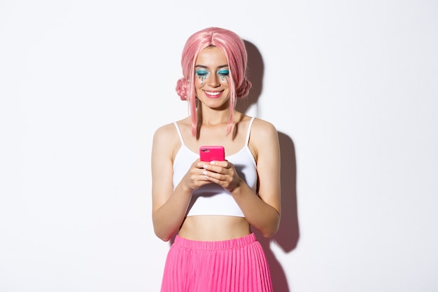 Retrato de uma linda mulher com peruca rosa anime e maquiagem brilhante, vestida para festa, sorrindo e olhando para o celular, em pé.