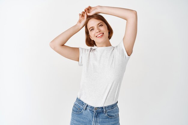 Retrato de uma linda mulher feliz com maquiagem de luz natural, descansando as mãos na cabeça e sorrindo, em pé de jeans e camiseta na parede branca