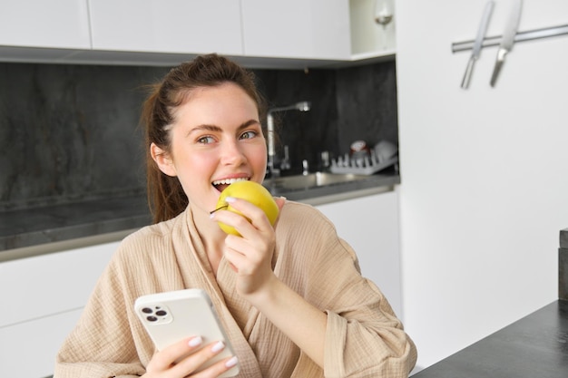 Foto grátis retrato de uma linda mulher feliz sorrindo comendo uma maçã na cozinha, sentada em casa, de roupão de banho