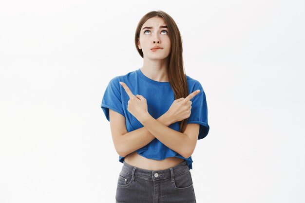 Retrato de uma linda mulher insegura em uma camiseta azul mordendo o lábio olhando para cima insegura cruzando as mãos contra o corpo apontando para a esquerda e para a direita sobre a parede cinza