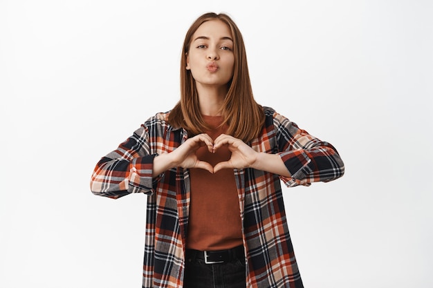 Foto grátis retrato de uma linda mulher loira beijando, mostrar sinal de amor de coração, como alguém, clima de encontro romântico, simpatia por alguém, de pé apaixonado e terno contra uma parede branca