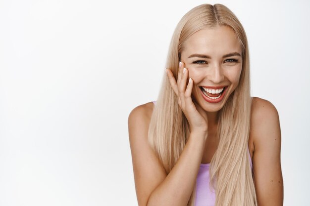 Retrato de uma linda mulher loira sorrindo rindo e tocando a pele facial macia e brilhante em pé sobre fundo branco Copiar espaço