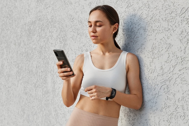 Retrato de uma linda mulher morena com blusa esportiva branca segurando um telefone inteligente