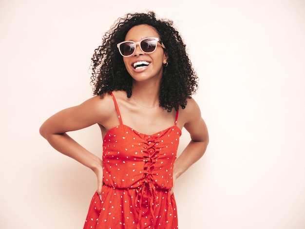 Retrato de uma linda mulher negra com penteado de cachos afro Modelo sorridente vestido com vestido vermelho de verão Mulher despreocupada sexy isolada parede branca em estúdio em óculos de sol