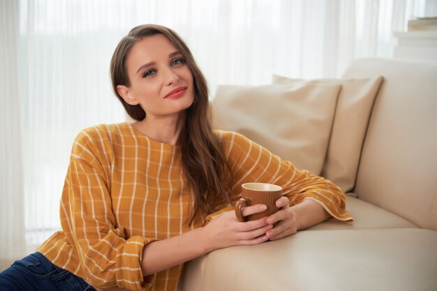 Retrato de uma linda mulher sentada no chão no sofá tomando chá quente e sorrindo