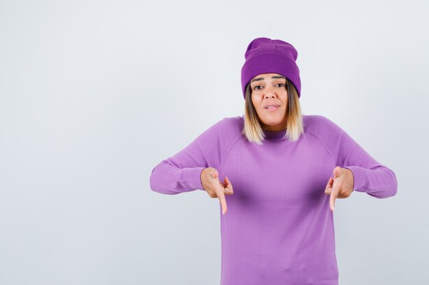 Retrato de uma linda senhora apontando para baixo com um suéter, um gorro e olhando confiante para a frente