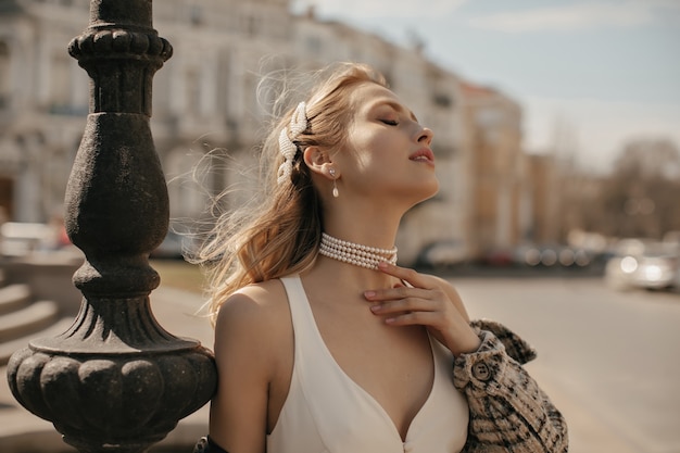 Foto grátis retrato de uma linda senhora loira em um elegante vestido de seda branca, jaqueta xadrez e colar de pérolas tocando suavemente o pescoço e posando na praça da cidade