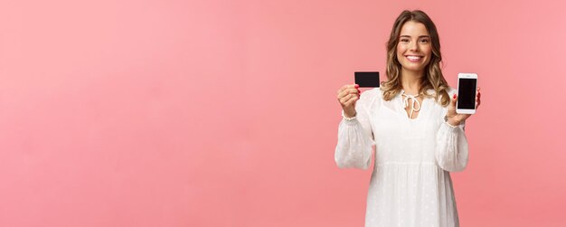 Retrato de uma loira bonita satisfeita em vestido branco, mostrando seu cartão de crédito e aplicativo de telefone celular, aplicativo de compras on-line ou site de banco sorrindo câmera em pé fundo rosa