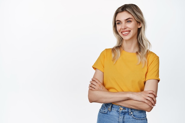 Retrato de uma loira confiante sorrindo de braços cruzados no peito e olhando de lado em camiseta amarela e jeans fundo branco