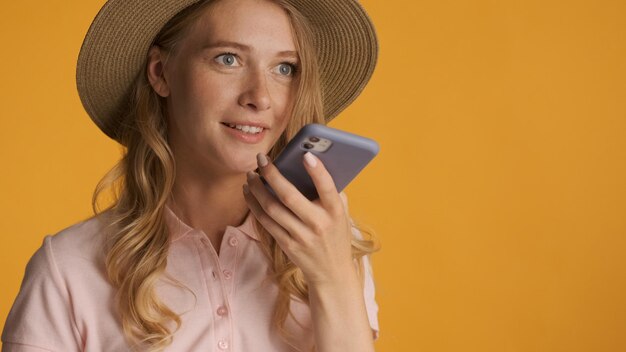 Retrato de uma loira excitada de chapéu usando gravador de comando de voz no smartphone sobre fundo amarelo