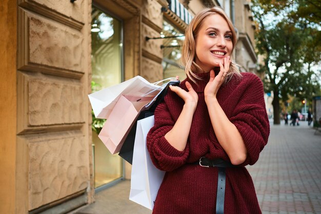 Retrato de uma loira sorridente atraente com sacolas de compras olhando alegremente para longe na rua da cidade