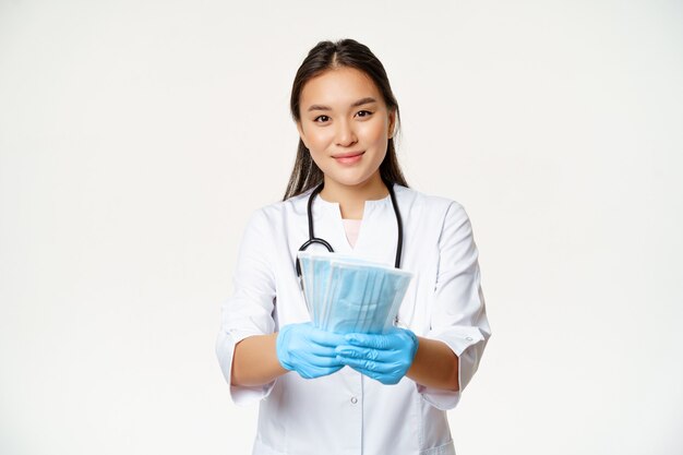 Retrato de uma médica asiática, enfermeira em luvas de borracha, dando máscaras médicas estéreis e sorrindo, medidas para prevenir a doença de coronavírus, fundo branco.